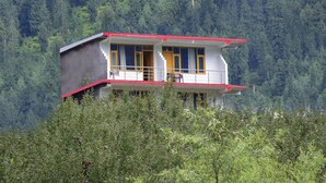 A two-story building with a red roof and white walls, surrounded by dense forest. - Madhuvan 2BHK, W/ Food, parking, Mountain View (Manali)