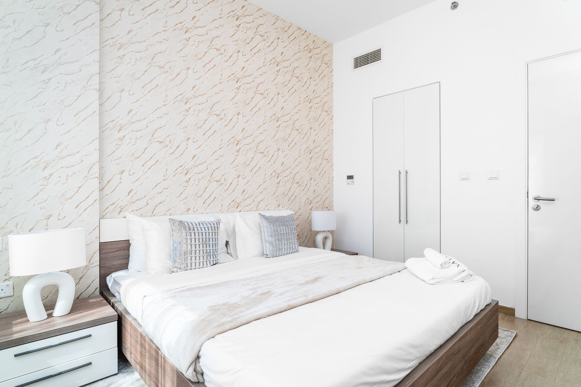 A modern bedroom with a marble-patterned wall, a bed with white bedding, a bedside table with a lamp, and a wardrobe.