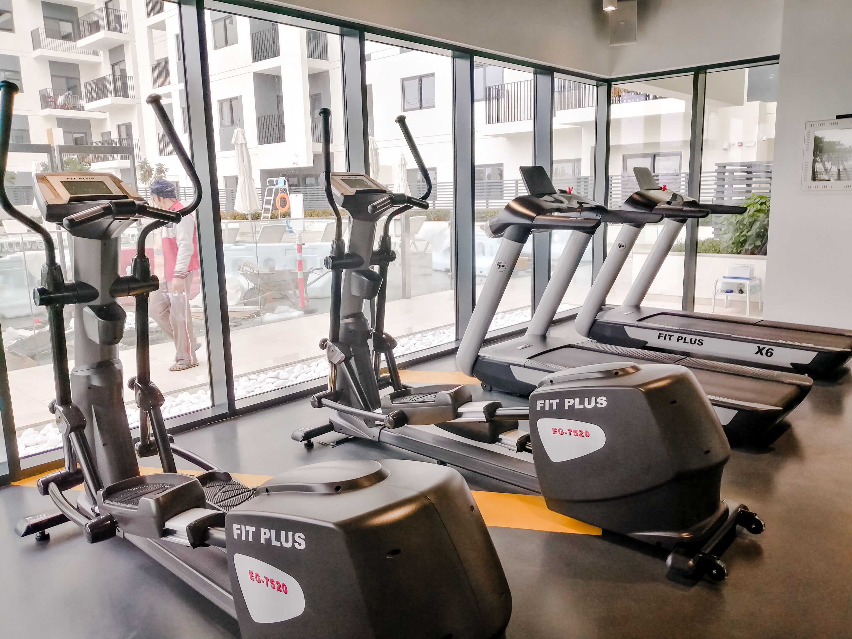 A gym with various cardio machines, large windows, and a view of a residential building.