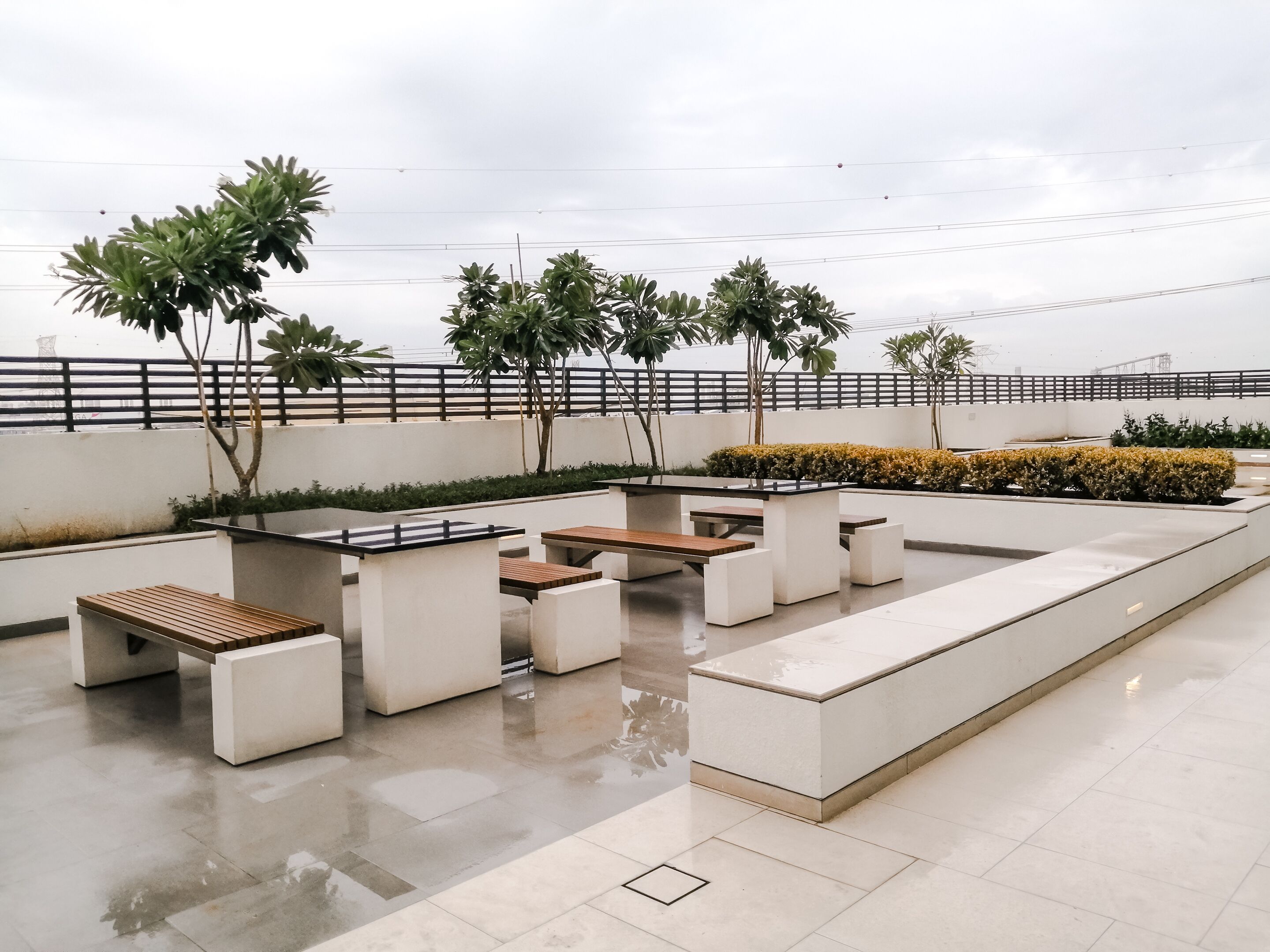 An outdoor seating area with concrete tables and benches, surrounded by greenery and a white fence.