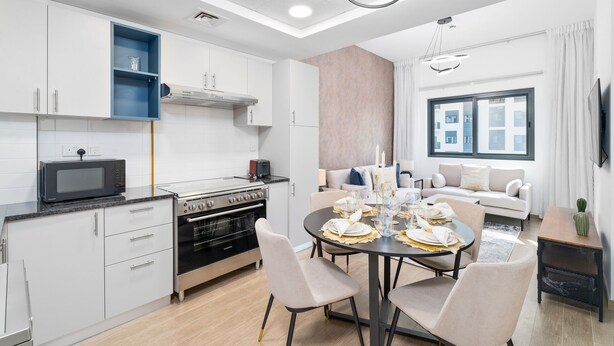 Een moderne keuken met witte kasten, een ingebouwde oven en een magnetron. Er is een eethoek met een ronde tafel voor vier personen, en een bank in de woonkamer.