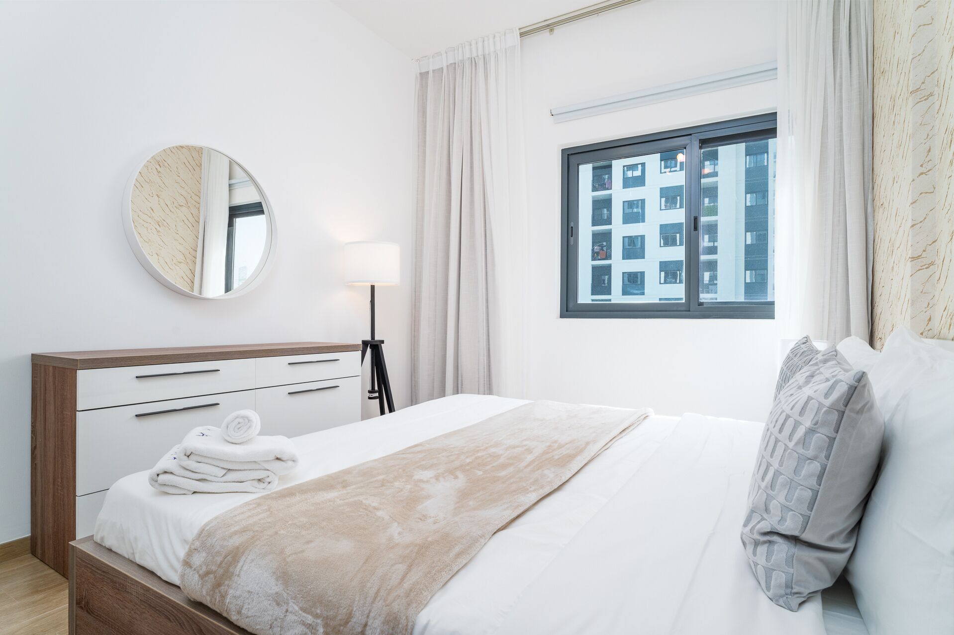A modern bedroom with a bed, a bedside table with a lamp, a round mirror, and a window with a view of buildings.
