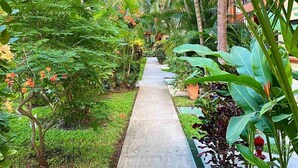 Un chemin en béton, bordé d’une végétation luxuriante et de plantes en fleurs, mène à un bâtiment doté de balcons.