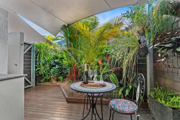 Une terrasse avec une table ronde et des chaises, entourée de différentes plantes et d’un petit espace aménagé en jardin.