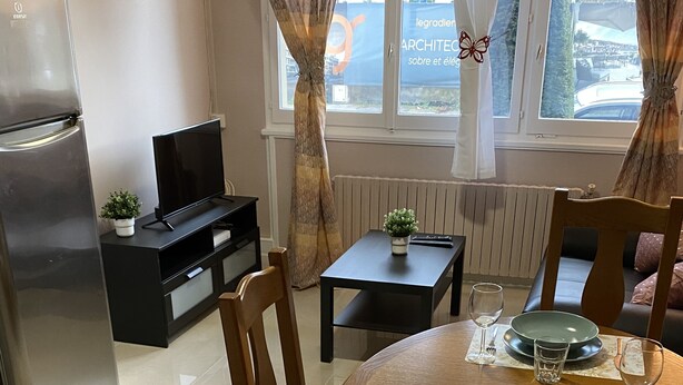 A dining area with a round table set for four, a black TV stand, a refrigerator, and a window with curtains. - LES IRIS ANNEMASSE (Annemasse)