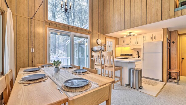 A modern kitchen with wooden cabinetry, a dining area with a wooden table set for four, and a view of the outdoors through large windows.