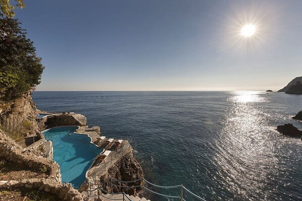 Ein Poolbereich mit Stufen zum Wasser, umgeben von felsigem Gelände und freiem Blick auf das Meer.
