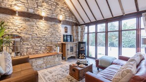 Un salon chaleureux aux murs en pierre, aux poutres apparentes et aux grandes fenêtres qui offrent une vue sur l’extérieur.