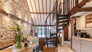 Un intérieur rustique avec un mur en pierre, des poutres apparentes, un escalier en colimaçon, un coin repas avec une table dressée pour deux personnes et une cuisine équipée d’appareils modernes.