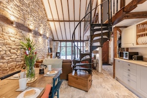 A rustic interior with a stone wall, wooden beams, a spiral staircase, a dining area with a table set for two, and a kitchen with modern appliances.