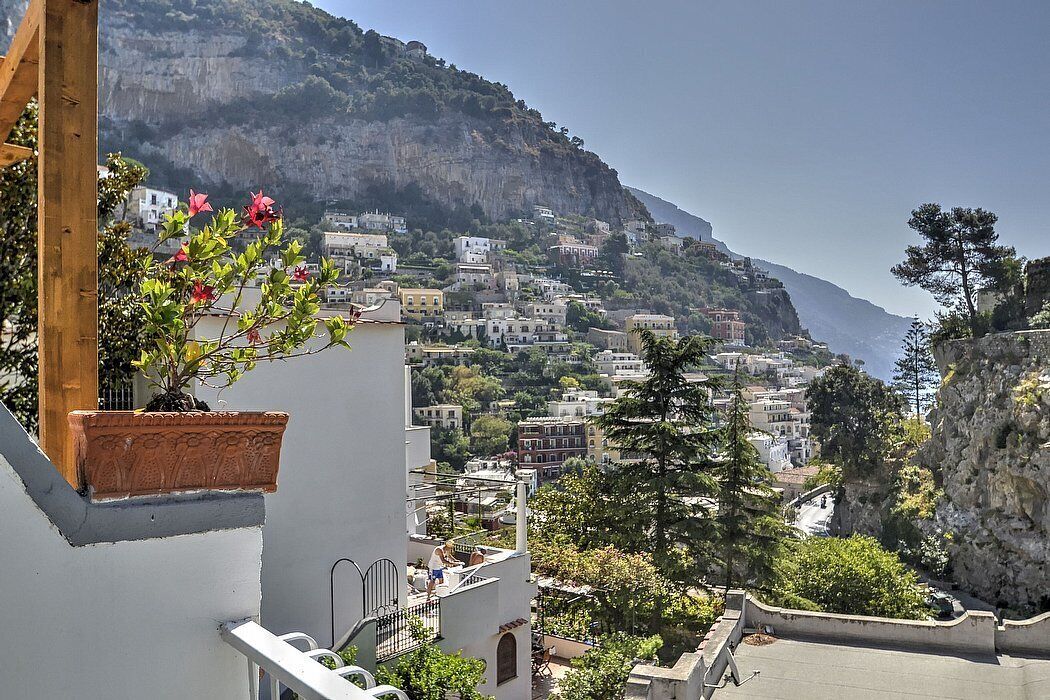 A coastal town with buildings on a hillside, a balcony with a potted plant, and a clear view of the sea.