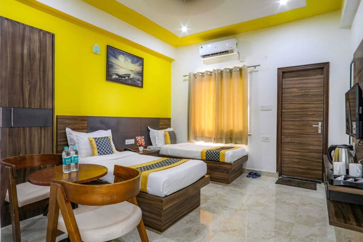 A hotel room with two beds, a dining table, chairs, a TV, and an air conditioning unit.
