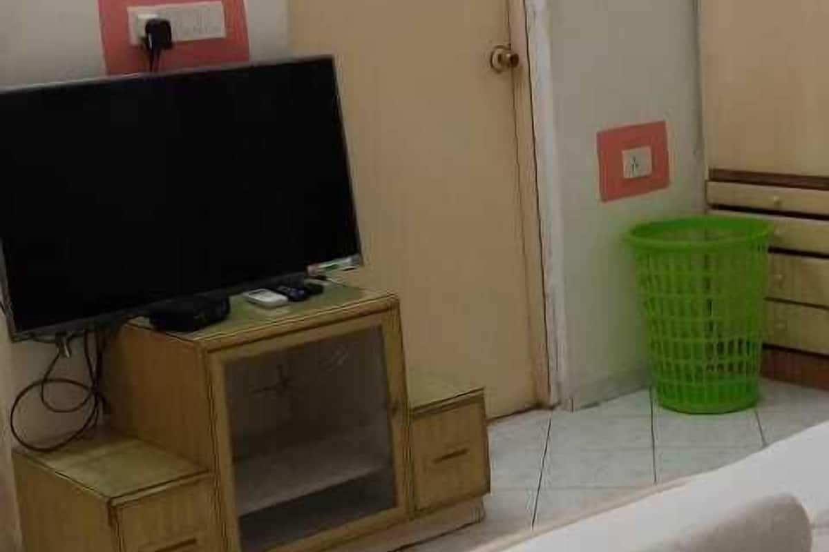A hotel room with a bed, a television on a wooden stand, a green basket, and a door with a light switch.