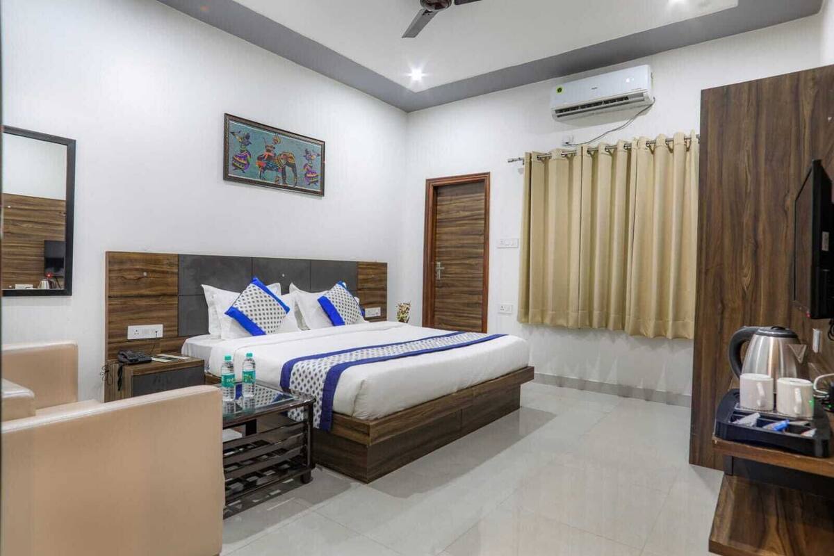 A hotel room with a bed, bedside tables, a mirror, a painting, an air conditioning unit, a window with curtains, and a coffee maker.