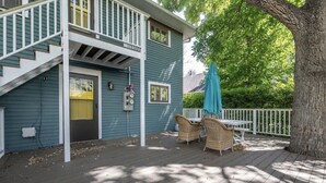 A two-story house with a deck, a covered patio area with wicker furniture, and a large tree.