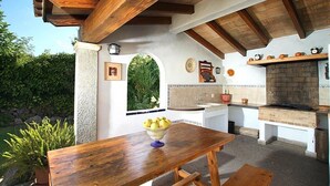 An outdoor kitchen with a brick oven, white cabinetry, and a wooden table with a bowl of fruit. - CAN VILA with pool, A/C, Wifi, close to the beach, table tennis, outdoor barbecue (IB)