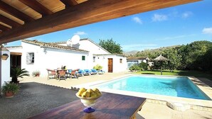 A pool area with a wooden pergola overhead, a dining table with a bowl of fruit, and a white building with a tiled roof in the background. - CAN VILA with pool, A/C, Wifi, close to the beach, table tennis, outdoor barbecue (IB)