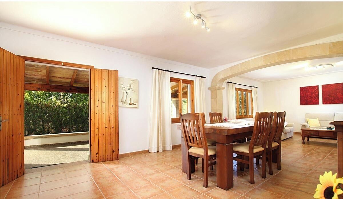 A dining area with a wooden table and chairs, a sofa, and a view of greenery through large windows.