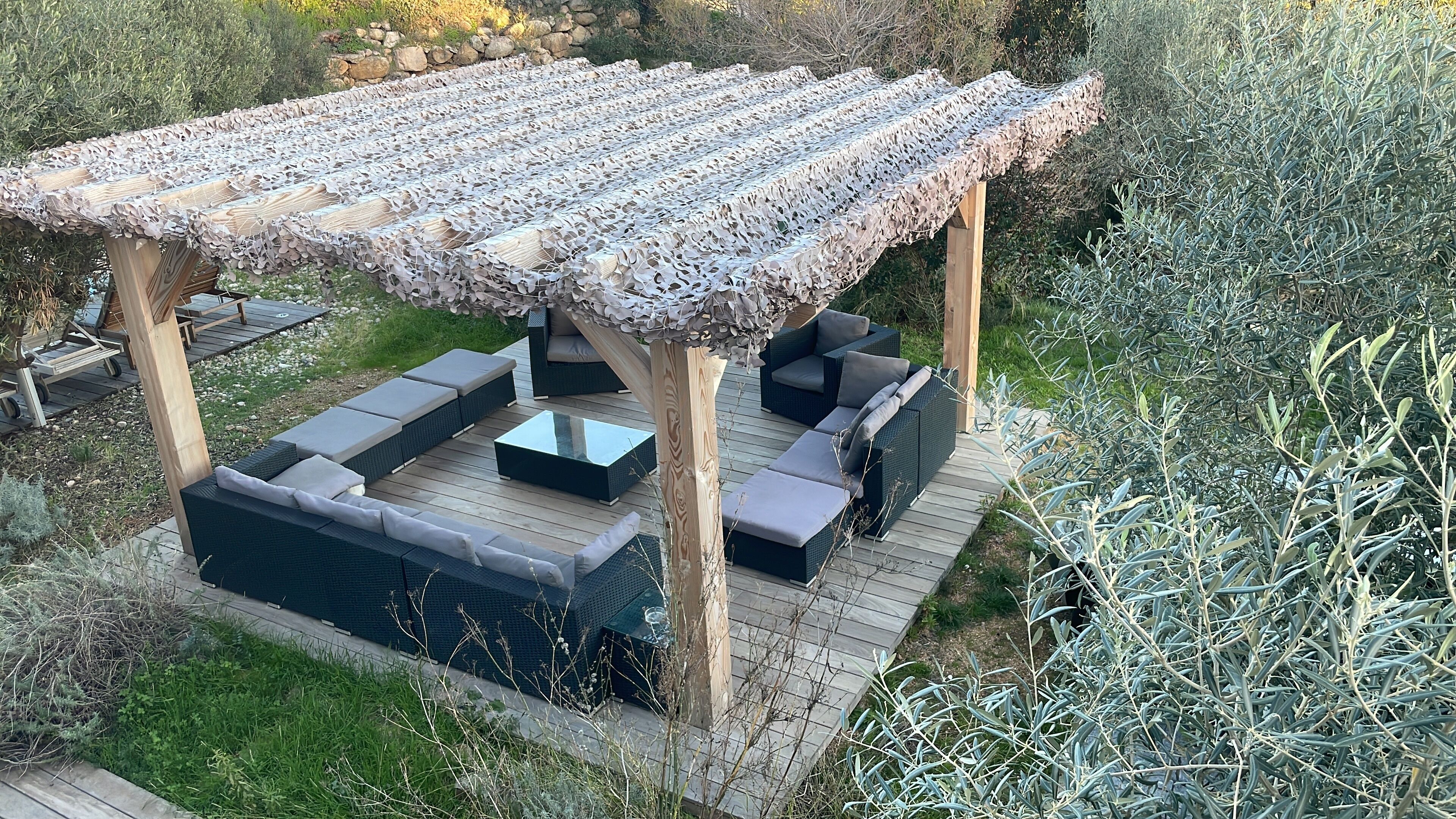Une pergola en bois avec un toit orné de motifs, un canapé intégré et une table basse en verre sur une terrasse en bois.