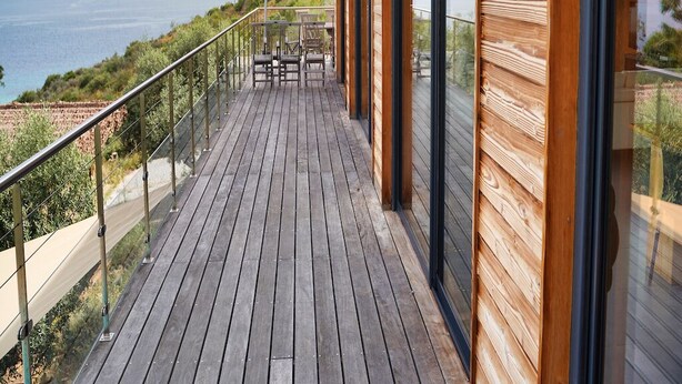 Une terrasse en bois dotée d’une balustrade en verre, offrant une vue sur un paysage côtier.