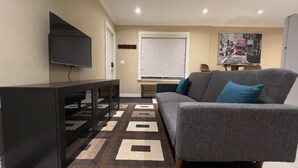 Een woonkamer met een grijze bank, een flatscreen-tv aan de muur, een kamerkleed met een patroon en een raam met zonwering.