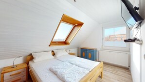 Ein Schlafzimmer mit schrägem Dach, einem Oberlicht, einem Bett mit weißer Bettwäsche, einem hölzernen Nachttisch, einem blauen Schrank und einer Heizung.