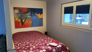 A bedroom with a bed, a window with blinds, a painting on the wall, and a view of the outside. - Modern studio in 1970s house in Haslital (Schattenhalb)