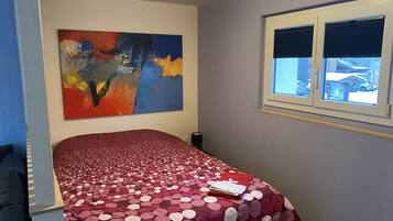 Une chambre à coucher avec un lit, une fenêtre avec des stores, un tableau au mur et une vue sur l’extérieur.