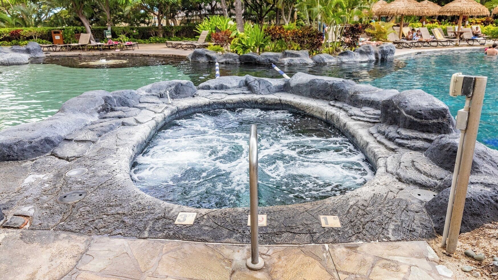A hot spring surrounded by rocks and a pool area with lounge chairs and umbrellas.