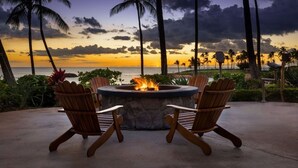 A fire pit with four wooden chairs around it, overlooking a beach with palm trees and a sunset. - Ko Olina | Beach & Pool Access | Tropical Resort Stay (Kapolei)