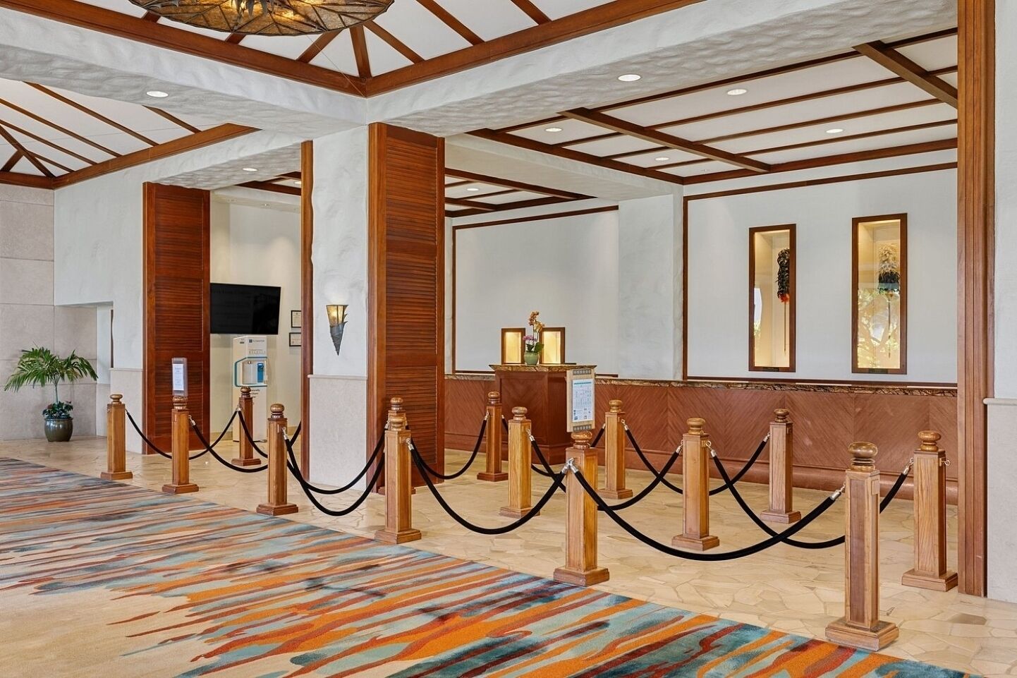 Un vestíbulo de hotel con mostrador de recepción, columnas de madera, alfombra estampada y techo con vigas a la vista.