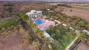 Vista aérea de un hotel con piscina, rodeado de jardines ajardinados y palmeras.