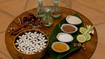 A wooden tray with various spices and ingredients, including a bowl of white flowers, cinnamon sticks, cloves, and green leaves.