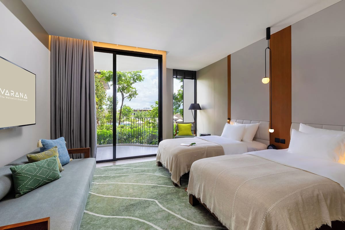 A modern hotel room with two beds, a sofa, a large window with a view of greenery, and a flat-screen TV mounted on the wall.