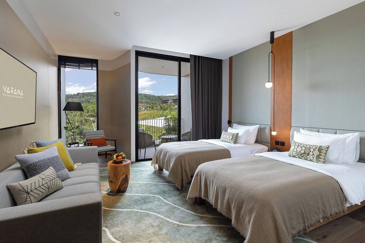 A modern hotel room with two beds, a sofa, a small table with fruit, and a balcony with a view.