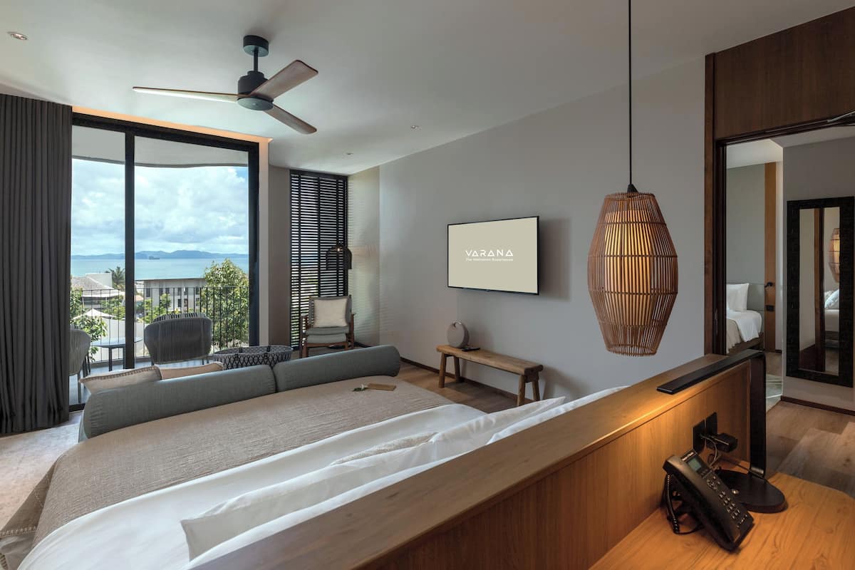 A modern hotel room with a large bed, a desk, a chair, a standing lamp, and a view of the sea.