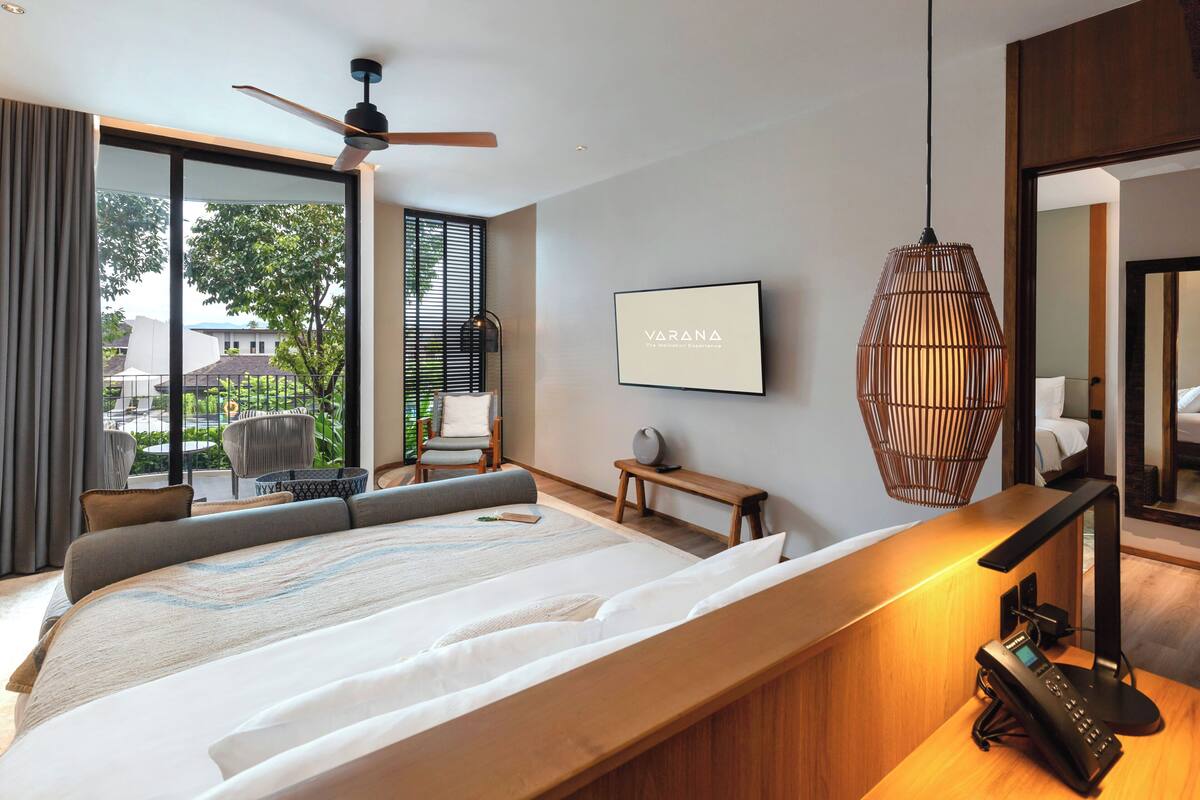 A modern hotel room with a large bed, a ceiling fan, a large window with a view of the outdoors, a mounted TV, and a bedside table with a lamp.