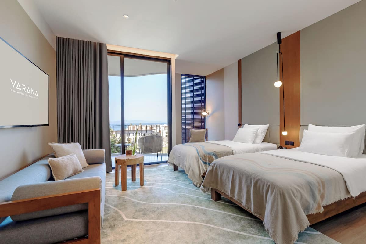 A modern hotel room with a large bed, a sofa, a small table, and a balcony with a view.