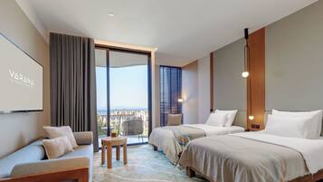 A modern hotel room with a large bed, a sofa, a small table, and a balcony with a view.