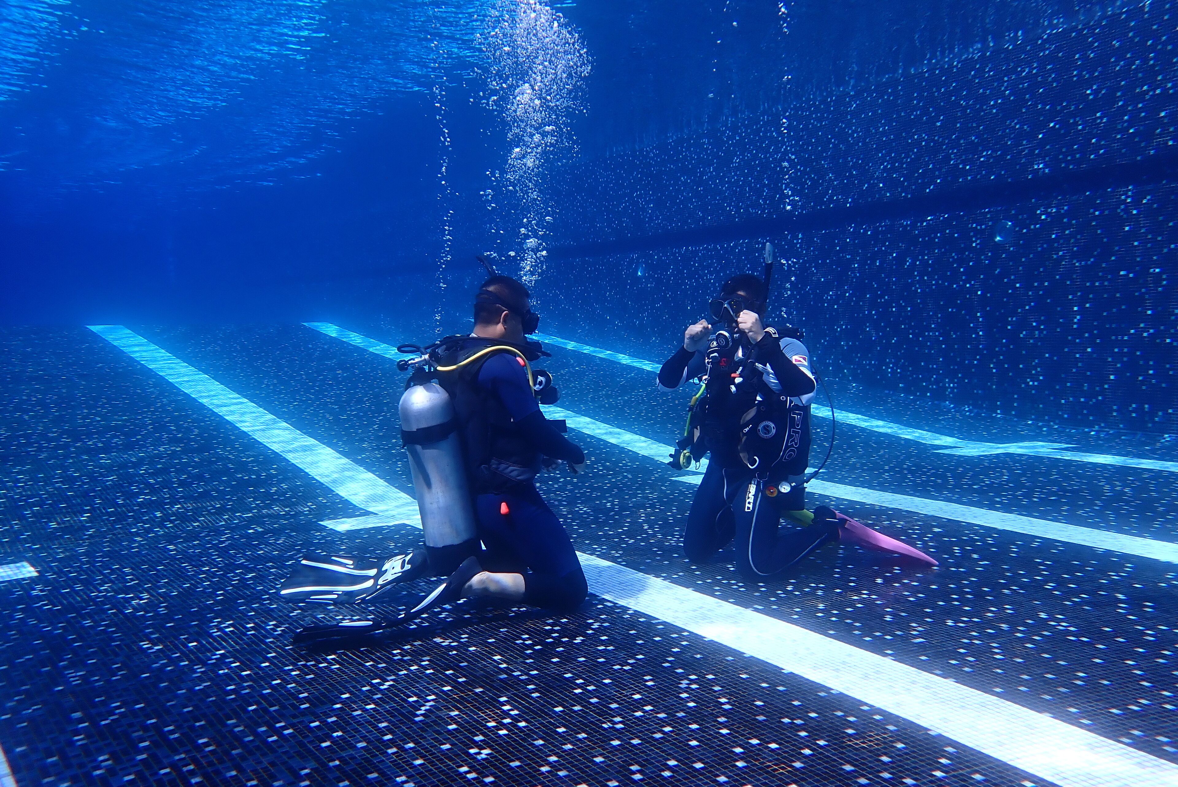 Two individuals in scuba diving gear on a submerged platform with a blue-lit background.