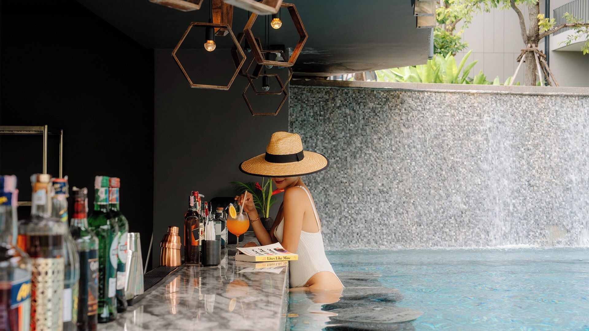 A person in a white swimsuit at a poolside bar with various bottles and a magazine.