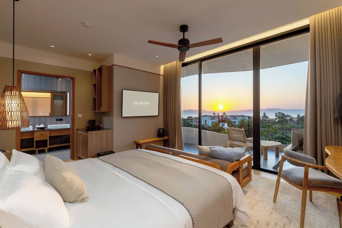 A modern hotel room with a large bed, a desk, a chair, and a balcony with a city view.