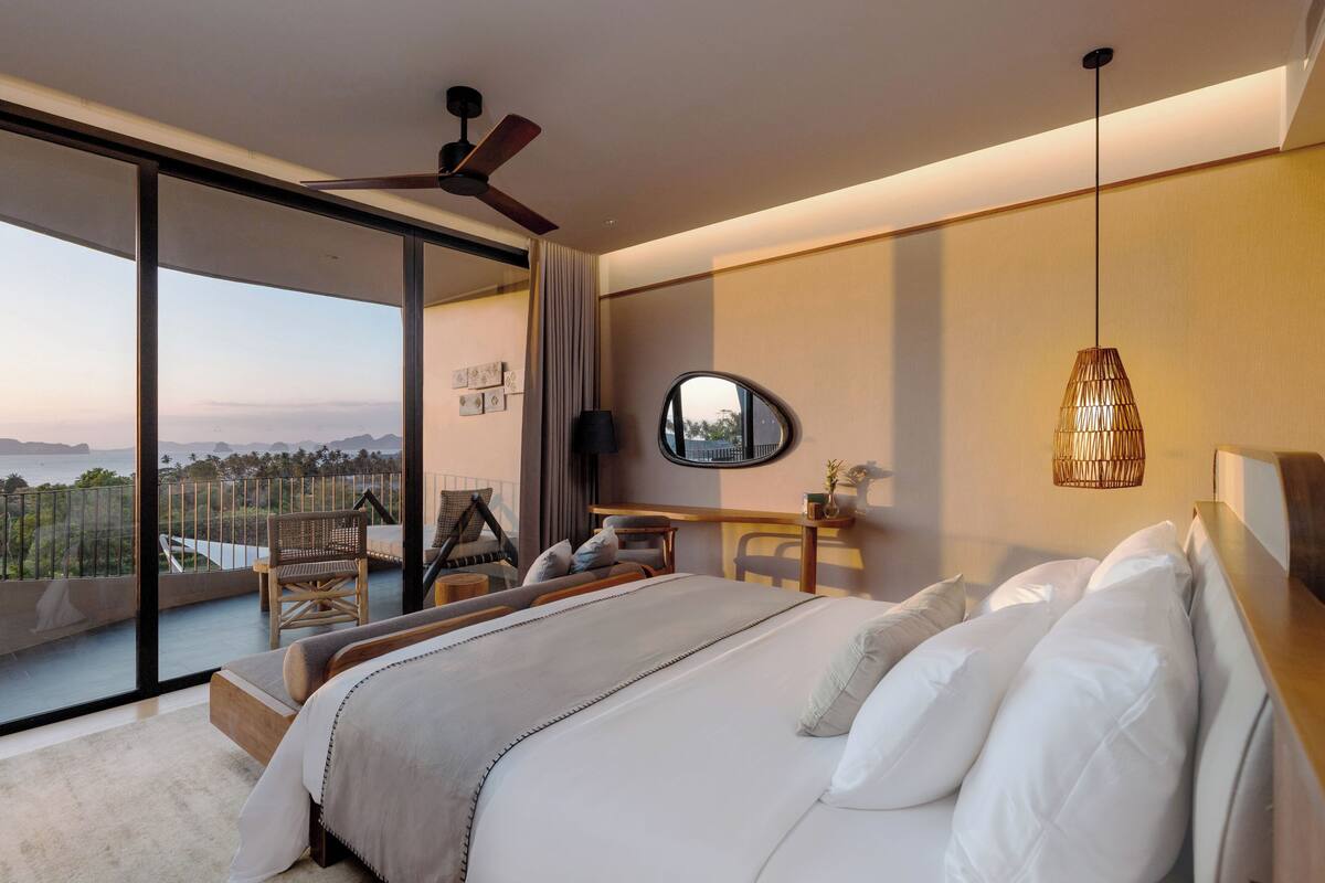 A modern hotel room with a large bed, a ceiling fan, a view of the ocean, and a balcony with a chair.