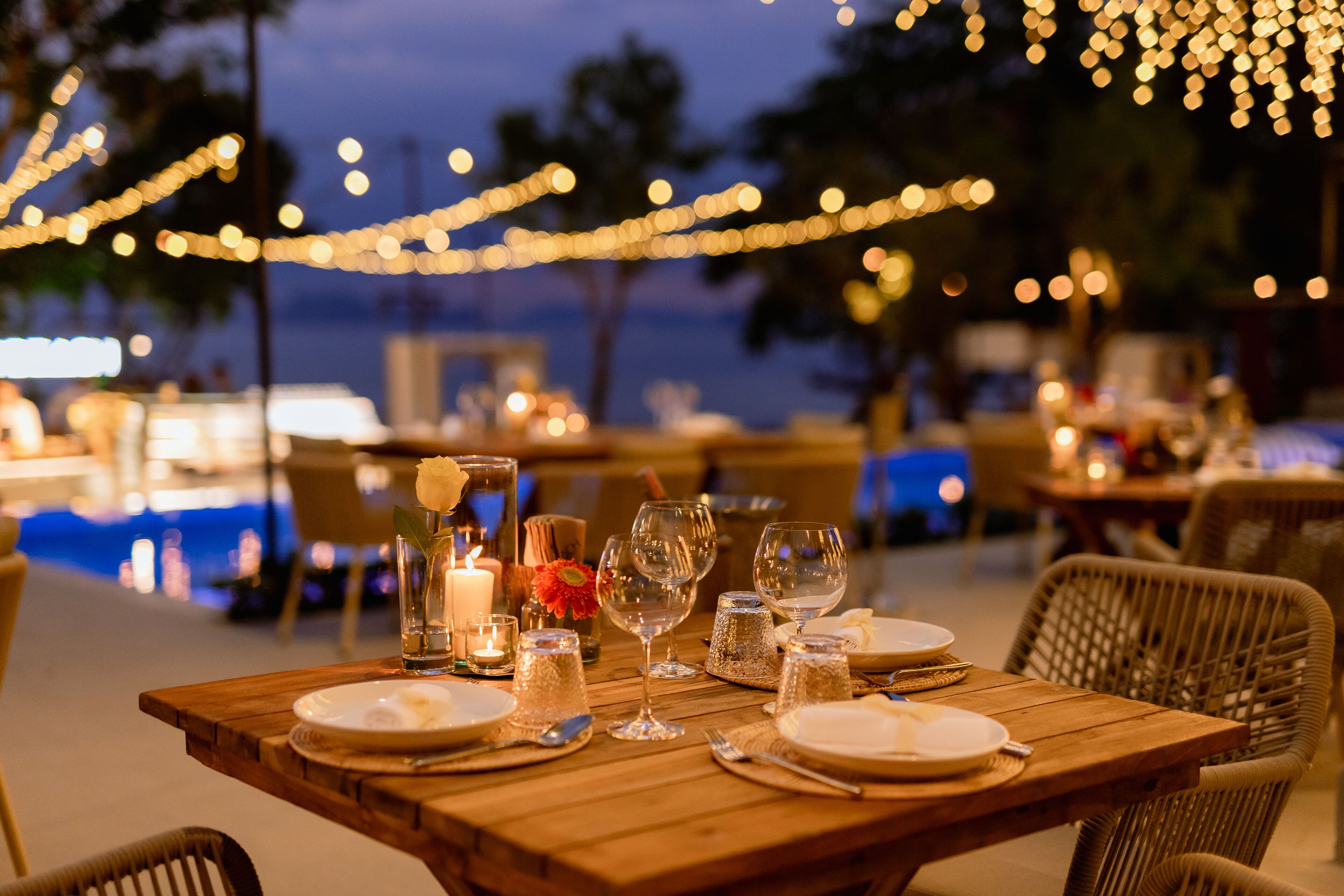 An outdoor dining area with tables set for a meal, string lights overhead, and a view of the ocean in the background.