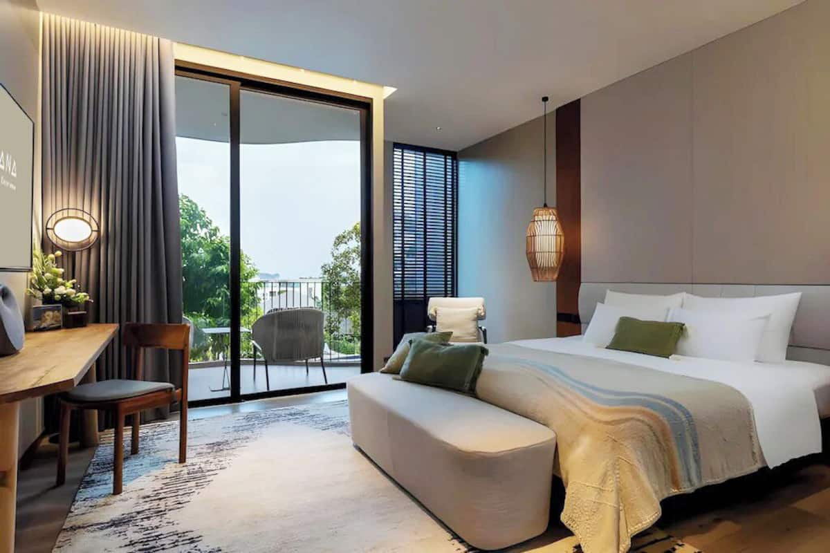 A modern hotel room with a large bed, a desk, and a chair. There is a balcony with a view of the outdoors.