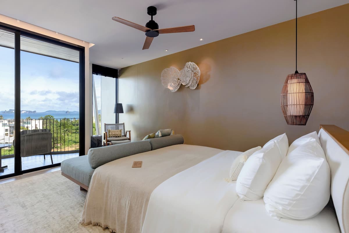A modern bedroom with a large bed, a ceiling fan, a view of the city, and a balcony with a chair.