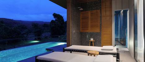 A modern spa room with two massage tables, a wooden ceiling, a glass shower enclosure, and a view of a pool at dusk.