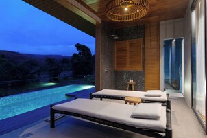 Un spa moderno con dos camillas de masaje, techo de madera, cabina de ducha de vidrio y vista a una piscina al atardecer.