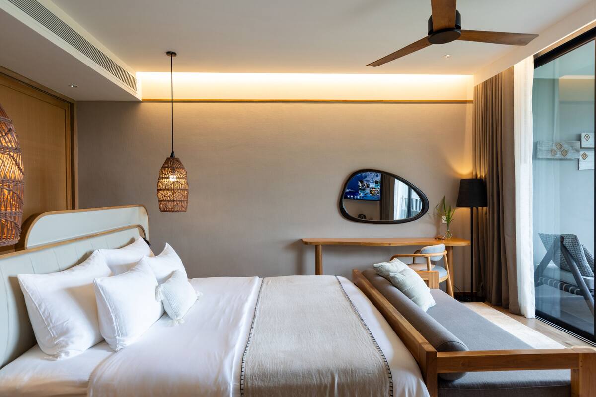 A hotel room with a bed, a desk, a chair, a ceiling fan, and a large window.