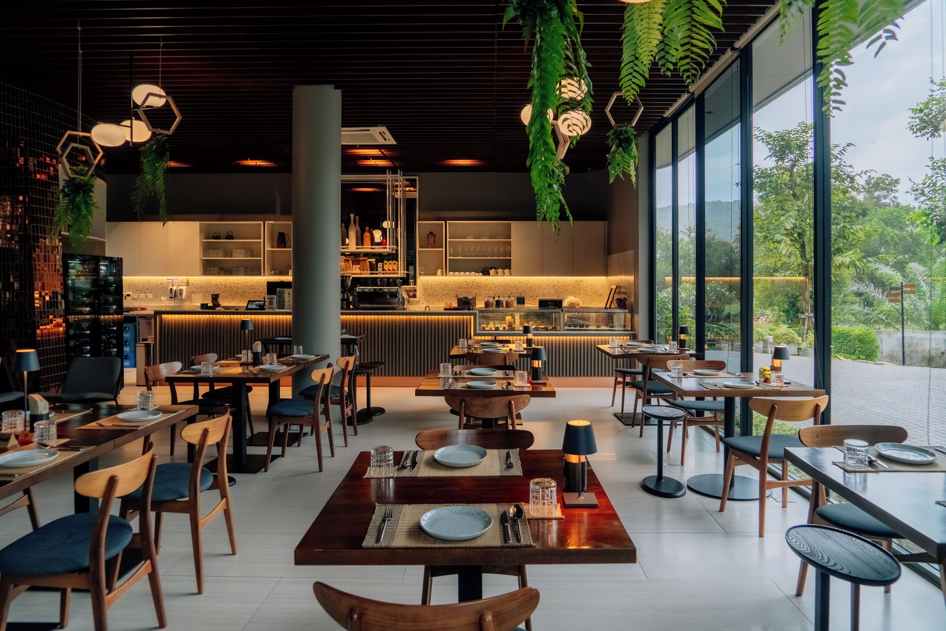 Een modern restaurant met houten tafels en stoelen, een bar en grote ramen met uitzicht op groen.